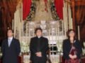 Celebración religiosa con la imagen de la Virgen en un altar ornado con velas y flores, acompañada por personas en trajes formales.