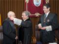 Tres hombres en trajes formales, uno de ellos recibiendo una medalla. En el fondo, un escudo y bandera nacional. Sede de gobierno o institución oficial.