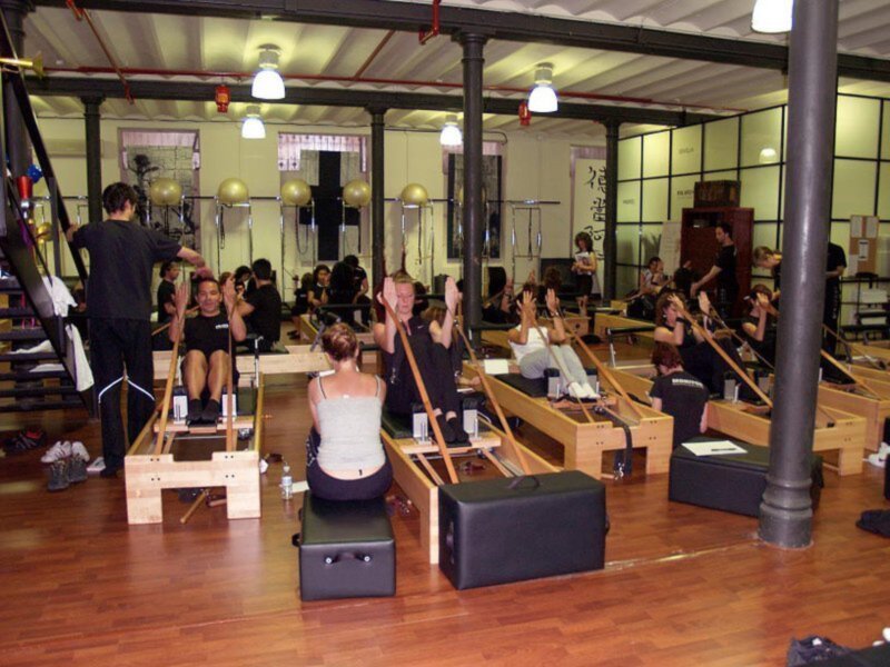 Una clase de pilates en un gimnasio con personas sentadas en máquina de remo, levantando las manos. El ambiente es acogedor con iluminación natural y estructuras industriales en el fondo.