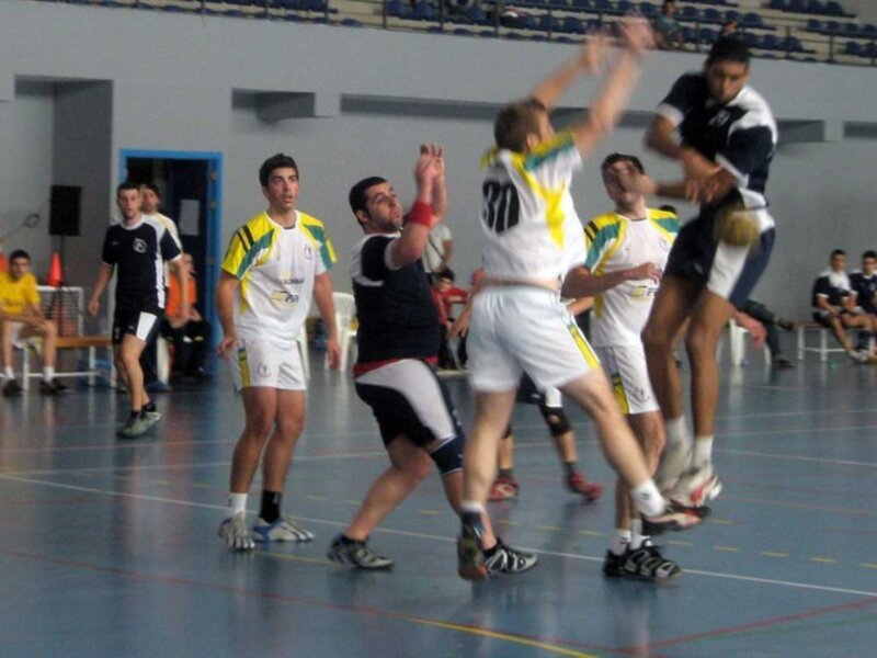 Un equipo de baloncesto en acción durante un partido. Los jugadores están en una cancha azul con asientos en el fondo. Un jugador de blanco y amarillo se levanta para lanzar la pelota, mientras otros jugadores intentan bloquear o defender. La escena muestra una competición intensa en un gimnasio con asientos y equipos alrededor.