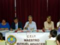 Una reunión formal con cuatro personas sentadas en una mesa, uno de ellos hablando en un micrófono, frente a un fondo de cortinas rojas y una bandera de CEIP Maestro Manuel Aparcercer.