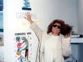 A una mujer con gafas de sol, posando frente a un cartel promocional del Carnaval 1997 en Chipiona, con una ilustración de un personaje y la fecha del evento.