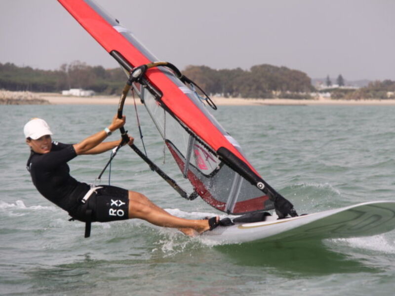 Un windsurfer en acción, con una vela roja y negra, navega sobre el agua azul. La persona está en una tabla de windsurf con un casco blanco y un traje negro, controlando la vela con una barra. El fondo muestra una costa con árboles y un edificio blanco, bajo un cielo despejado.