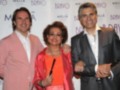 Tres personas sonrientes posan juntas en un evento con un fondo blanco que contiene el logo de Meliá y Bravo. La persona central lleva una chaqueta roja brillante, mientras que los otros dos están con trajes formales.