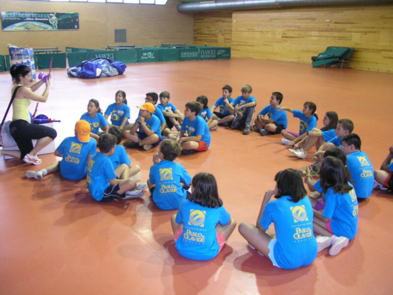 Niños sentados en círculo, con camisetas azules y gorros amarillos. Una persona en la izquierda sostiene un objeto rojo. Fondo de una sala de deportes con paredes verdes y amarillas, un techo inclinado y una manta verde en el fondo.