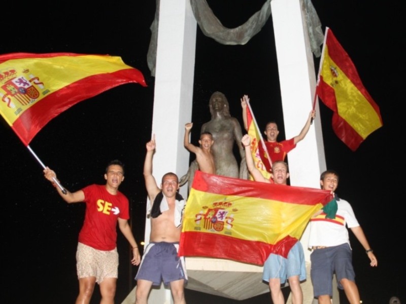 Jóvenes celebrando con banderas españolas en una escultura al atardecer.