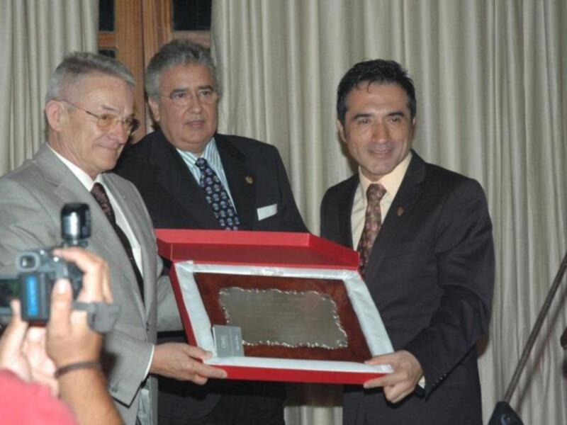 Un hombre en un traje gris sostiene una caja roja con un trozo de metal plateado, mientras dos hombres en trajes de negocios observan. La escena está ambientada en un entorno formal, posiblemente una ceremonia o evento profesional.