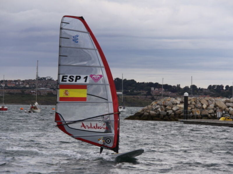 Una persona realiza windsurfing en el agua con una vela roja y blanca que muestra la bandera de España. En el fondo, se pueden ver otras embarcaciones y una costa con rocas.