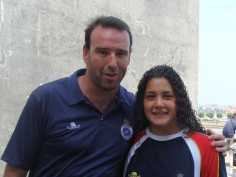 Dos personas sonrientes, uno con una camiseta azul y otro con una camiseta multicolor. Fondo de una estructura de piedra y cielo azul.