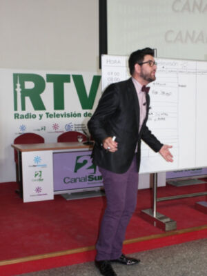 Presentador en una conferencia de radio y televisión, con un cartel de Canal Sur Televisión al fondo.