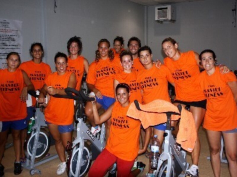 Un grupo de personas en una sala de fitness, todos vestidos con camisetas naranjas y pantalones cortos, posando para la foto. En el fondo, se pueden ver bicicletas de ejercicio y una pared con carteles de instrucciones.