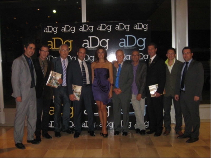 Una celebración empresarial con una mujer en el centro y varios hombres a su alrededor, todos vestidos formalmente, posando frente a un fondo con el logotipo 'aDg'.