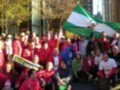 Una multitud de personas vestidas con chaquetas rojas y algunos con gorros, posando para una foto. Algunos sujetan banderas de diferentes países y un gran bandera verde, blanco y azul. En el fondo, se pueden ver edificios modernos con ventanas grandes.