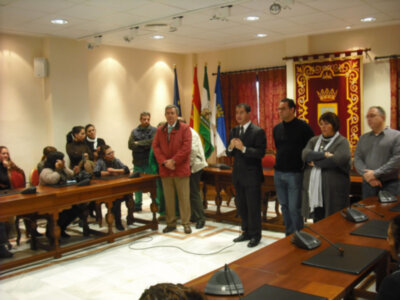 Un grupo de personas en una sala de conferencias, con un hombre en el centro hablando. Hay banderas y un escudo en la pared, una mesa con sillas al fondo y gente sentada.