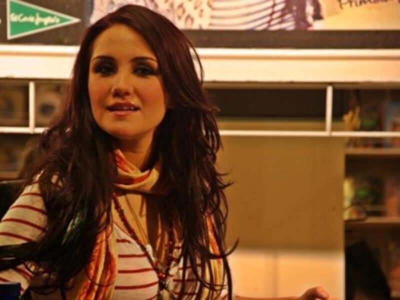 Una mujer con cabello largo y rizado, vistiendo una camiseta a rayas y una bufanda naranja, sentada en un entorno interior con estanterías de libros en el fondo.