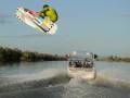 Un hombre realiza una maniobra de wakeboard en el aire, con un barco detrás que mueve la agua.