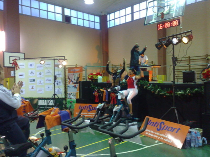 En una sala deportiva, un grupo de personas participa en actividades físicas. En el fondo, se puede ver una pizarra con información y un tablero electrónico que muestra la hora 16:00. La imagen también incluye elementos promocionales de IntiSport, como una camiseta y un balón.