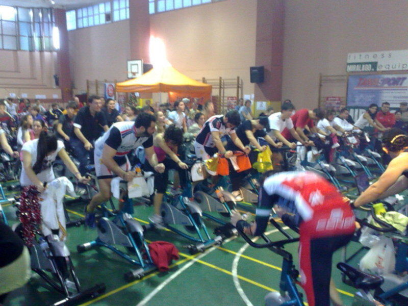Un grupo de personas en bicicletas estándar en una sala de fitness, con un ambiente animado y una iluminación brillante.