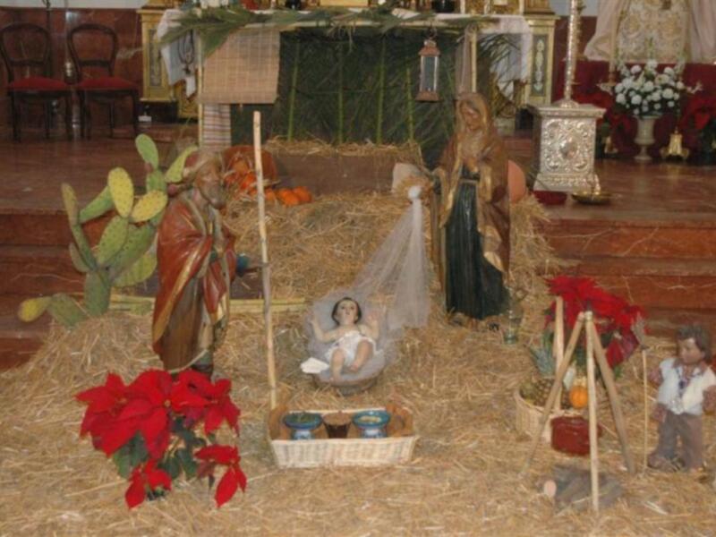 Escena navideña con un nacimiento en el centro, rodeado de adornos navideños como cactus y flores rojas. Una mujer y un hombre vestidos con trajes tradicionales están presentes, junto a un niño recién nacido en una hamaca. La escena está ambientada con esteras de paja y detalles navideños, creando un ambiente festivo.