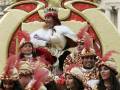 Imagen de un desfile navideño con personajes vestidos de reyes y reinas, rodeados por personas en trajes festivos con plumas.