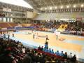 Un estadio deportivo con una cancha de baloncesto y voleibol, lleno de espectadores. La arena tiene gradas vacías en los costados y una barra blanca en el centro. La iluminación es intensa, destacando la actividad en el campo de juego.