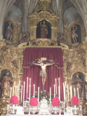 Altar con crucifijo central, velas y flores rosadas. Detalles dorados en el retablo.