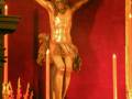 Una estatua de Cristo crucificado en una iglesia, rodeada por velas encendidas y flores rojas. El fondo es un muro rojo oscuro, iluminado por la luz de las velas.