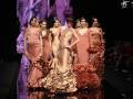 Modelos desfilan en la pasarela de moda, luciendo vestidos de colores pastel y con detalles elaborados. La escena es dramática, iluminada por una pantalla de fondo que refleja su imagen.