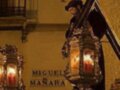 Imagen nocturna de una procesión religiosa en Semana Santa 2011, con un hombre vestido de negro cargando una cruz iluminada por dos faroles. La imagen muestra detalles como la decoración de la iglesia y el nombre "Miguel Manara" en una cruz.