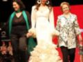 Una modelo camina por la pasarela con un vestido blanco encaje, acompañada de dos mujeres mayores. La escena se realiza en un escenario iluminado con luces y telones rojos.