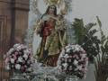 Estatua de la Virgen María con un niño en sus brazos, rodeada de flores y decoración. Fondo incluye una estructura arquitectónica y un cartel colgante.