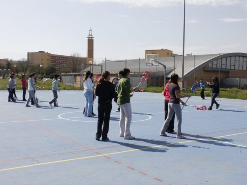 Una escena en un campo de baloncesto con varias personas jugando y observando. La imagen muestra a varios niños y adultos en una cancha de baloncesto, con algunos jugando con pelotas y otros observando. En el fondo se puede ver una torre de televisión y edificios a lo lejos. La cancha tiene líneas rojas y amarillas, y el cielo es claro con algunas nubes.