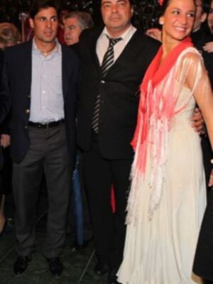 Una imagen de tres personas posando juntas, dos hombres y una mujer. La mujer lleva un vestido blanco con detalles en rojo y tiene el cabello recogido. Los hombres están con camisas y pantalones, uno de ellos con una corbata. La escena parece ser en un evento formal o ceremonial, posiblemente una fiesta o gala.