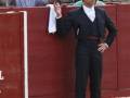 Un torero en el ruedo de una corrida de toros, con una camisa blanca, un chaleco y pantalones negros, mientras sostiene una botella de agua.