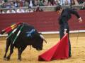 Un torero con un traje negro y una saya roja se enfrenta a un toro negro con cuernos blancos. El torero sostiene una saya roja y parece estar en acción, mientras el toro se mueve hacia él. La imagen muestra una escena típica de la tauromaquia en un anfiteatro lleno de espectadores.