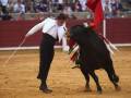 Un torero con un traje blanco y negro se enfrenta a un toro negro en una plaza de toros. El torero sostiene un bastón y una bandera roja con plumas blancas en su mano izquierda, mientras el toro se lanza hacia él. La arena es de tierra y la pared roja del fondo indica que se trata de una corrida de toros.