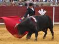 Un torero se enfrenta a un enorme toro negro en una plaza de toros. El torero sostiene una capa roja, mientras el toro avanza hacia él con fuerza. La arena es de tierra y la gente observa desde los muros alrededor.