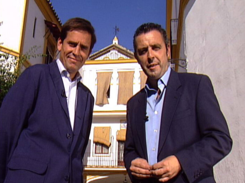 Dos hombres en trajes azules sonríen frente a una casa blanca con ventanas y un techo de tejas. El cielo es azul claro sin nubes.