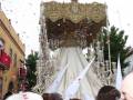 Fiesta religiosa con una imagen vestida de blanco y rodeada de banderolas blancas, mientras caen confeti rojo en el aire.