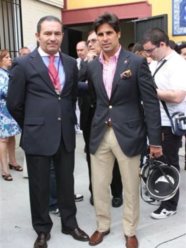 Dos hombres en trajes posan juntos frente a una fachada de edificio. Uno lleva un traje negro y corbata roja, mientras que el otro está con una chaqueta azul claro y pantalones beige. El hombre de la chaqueta tiene un bolso negro colgado del hombro y una gorra en el suelo. La escena parece ser un evento formal o ceremonial.