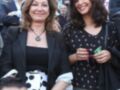 Dos mujeres sonrientes posan juntas en un evento público, con una de ellas sosteniendo un teléfono móvil. Ambas lucen elegantes y sonríen ampliamente, destacando su felicidad.