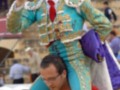 Un torero en traje tradicional sostiene un capote azul y dorado, mientras otro hombre lo lleva en sus hombros. La escena se desarrolla en un estadio con asientos vacíos y una torre de sonido visible.