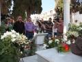 Una persona está enterrada en un cementerio, rodeada de flores blancas y rojas. Algunos asistentes están tomando fotos con cámaras digitales, mientras otros observan el momento. La escena es tranquila y triste, con un cielo despejado en el fondo.