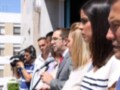 Una mujer con un vestido blanco y negro habla en una rueda de prensa, mientras otros asistentes observan.
