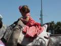 Una niña en un vestido rojo con lunares se sienta en el asiento de un caballo gris, con un arnés y una manta blanca debajo. El fondo muestra un cielo azul claro y algunos árboles en el horizonte.