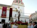 Una procesión religiosa en una calle con un paso de misterio amarillo, un altar con flores y un Cristo en la cruz. En el fondo, una torre de iglesia con banderas y un reloj.