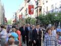 Una multitud camina por una calle con edificios históricos y banderas rojas. La gente está vestida formalmente, algunos con sombreros. El cielo es azul claro y el ambiente parece ser festivo o ceremonial.