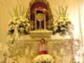 Un altar adornado con flores blancas y un ornamento dorado, en una habitación con cortinas amarillas.