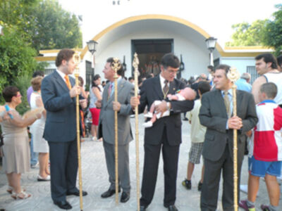 Un grupo de personas en trajes y sombreros participan en una ceremonia, sostienen palos y un bebé es sostenido por uno de ellos. La escena se desarrolla frente a una estructura con arco dorado y árboles al fondo.