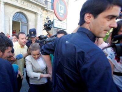 En un entorno urbano, una persona en el centro de la imagen está siendo filmada por varios fotógrafos y reporteros. La escena se desarrolla frente a un edificio con una gran puerta arqueada y un letrero que dice "CIRCUITO DE JUSTICIA". La atmósfera sugiere una situación de interés público, posiblemente relacionada con la justicia o un evento judicial.